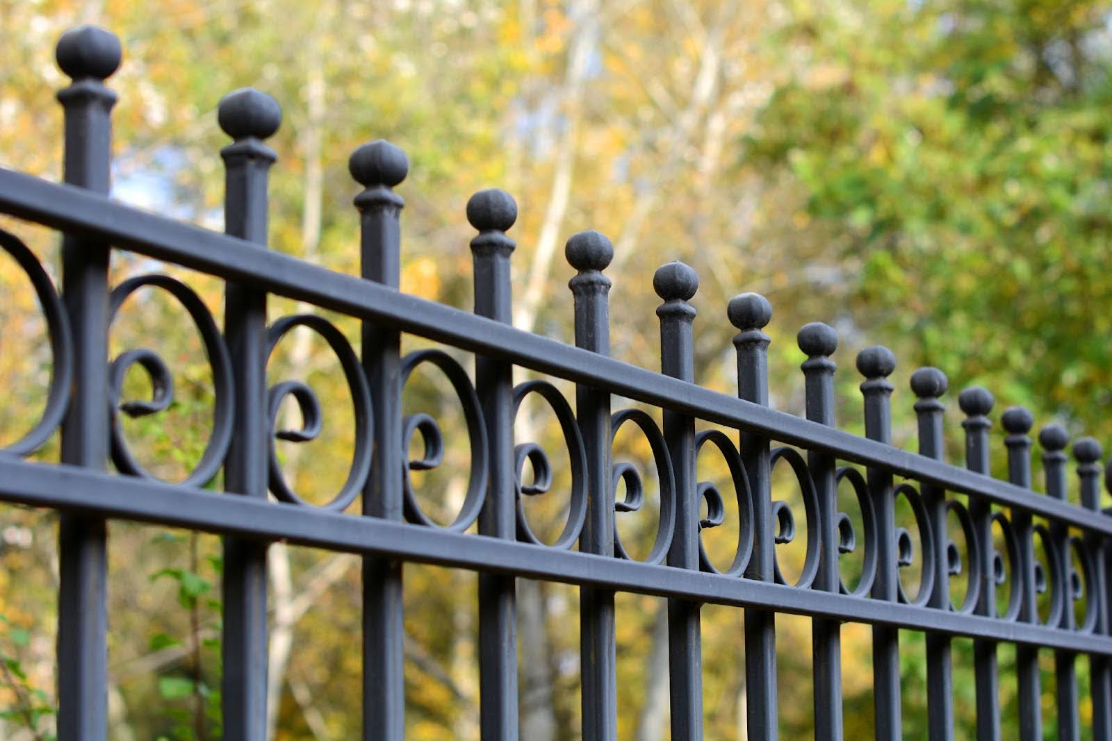 A decorative black wrought iron fence with circular patterns, set against a backdrop of trees.