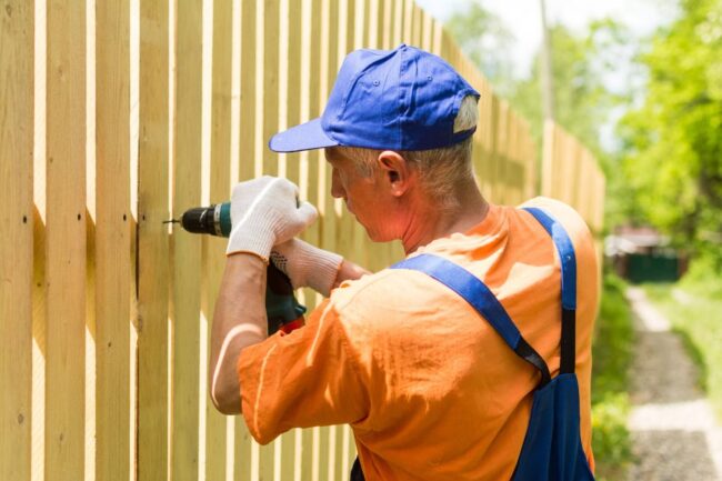 A worker in an orange shirt uses a power drill to install a wooden fence on a sunny day.